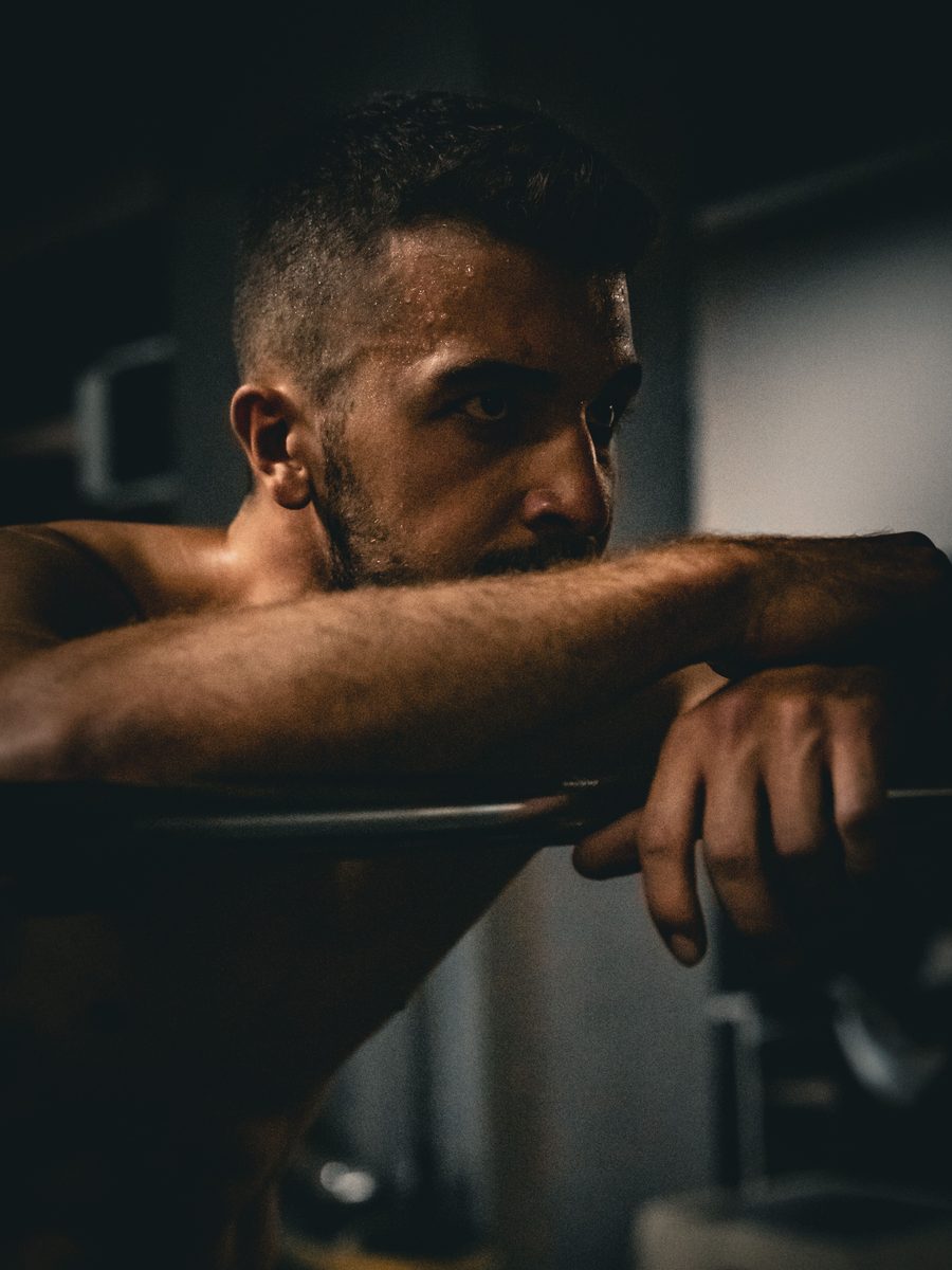 An athlete resting his arms on a barbell in a dim gym, looking off-frame