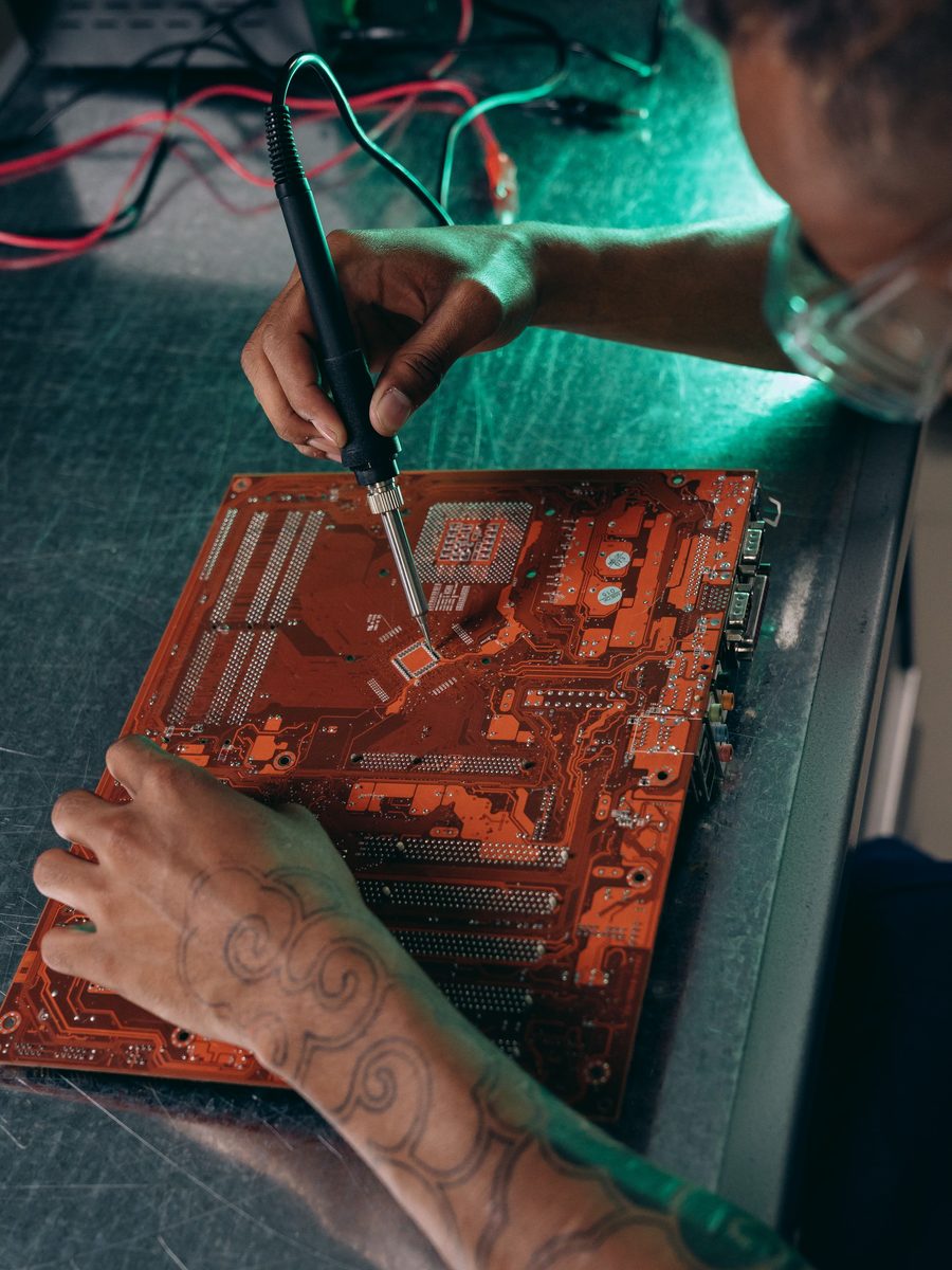 An engineer soldering a bare motherboard on a steel bench under green light